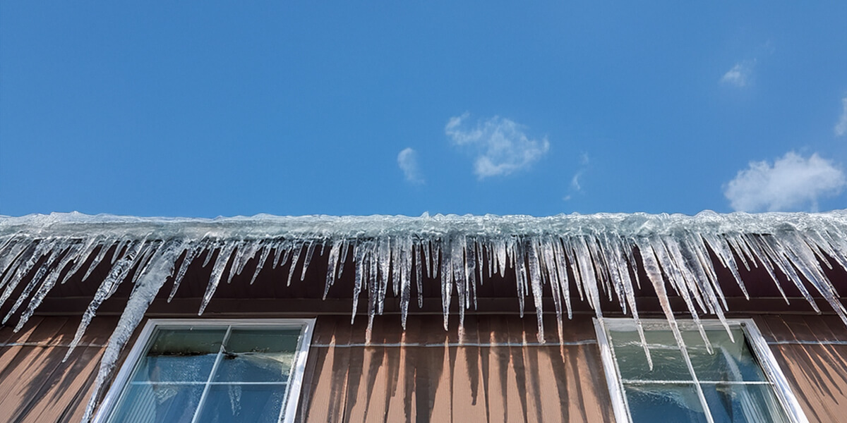 A large, thick sheet of ice hangs from the gutter of a house roof covered in snow, forming ice dams on the roof with icicles extending down to the ground. Snow piles up around the building on a bright winter day.
