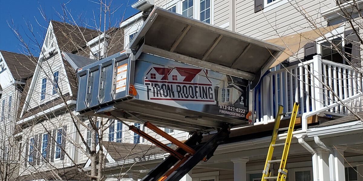 Une benne métallique identifiée Firon Roofing est soulevée par un treuil jusqu’à un étage supérieur d’une maison beige avec garnitures blanches. Une échelle jaune est appuyée contre le porche, et des branches d’arbres sans feuilles sont visibles.
