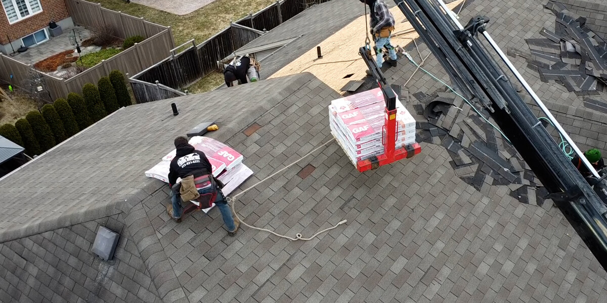 Un travailleur installe des bardeaux sur un toit en pente tandis qu’une grue soulève des paquets de bardeaux sur le toit. D’autres travailleurs et matériaux sont visibles, illustrant l’expertise des meilleurs couvreurs en Amérique du Nord dans un quartier résidentiel.