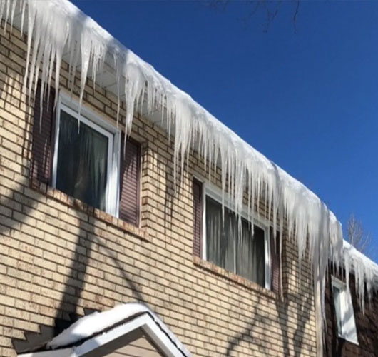 Maison en brique de deux étages avec de longs glaçons acérés suspendus au toit et des stores fermés aux fenêtres, sous un ciel bleu clair. La neige recouvre le toit inférieur, illustrant la nécessité d’un déneigement et d’un retrait de glace rapides.