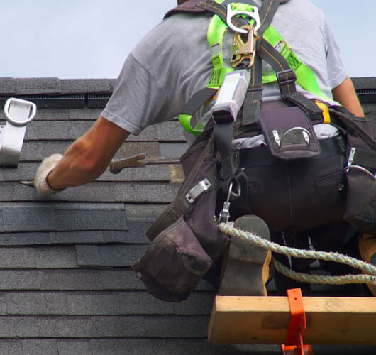 Un travailleur portant un équipement de sécurité et un harnais installe des bardeaux d’asphalte lors de réparations de toiture à Gatineau, utilisant un outil pour les fixer. Une corde de sécurité est attachée pour la protection contre les chutes.