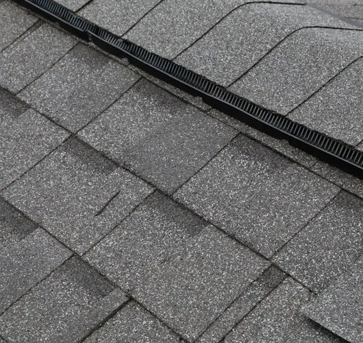 Vue rapprochée de bardeaux d’asphalte gris avec un évent faîtier noir courant le long du sommet, mettant en évidence la texture et le chevauchement des bardeaux tout en illustrant une ventilation efficace du toit.