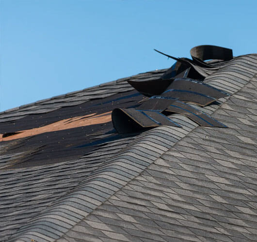 Toit endommagé avec plusieurs bardeaux d’asphalte soulevés, exposant la structure de bois en dessous, sous un ciel bleu clair. Les dommages causés par le vent sont évidents alors que les bardeaux semblent soulevés et nécessitent une réparation.