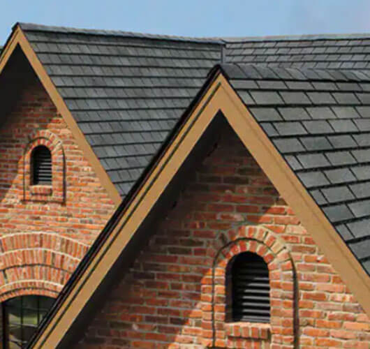Deux toits à pignons avec des bardeaux design gris et des murs extérieurs en brique rouge, chacun avec une lucarne en arc à persiennes. Le style architectural suggère un design classique ou traditionnel.