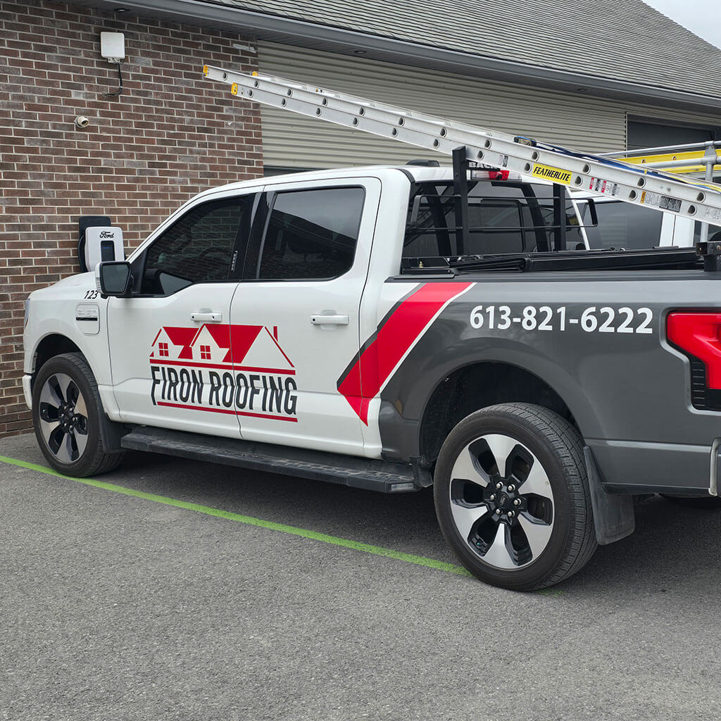 Une camionnette grise et blanche avec Firon Roofing et un numéro de téléphone imprimé sur le côté, garée à l'extérieur d'un bâtiment en briques avec des échelles dans la benne, prête à gérer toute réparation de toiture dont les clients d'Ottawa/Gatineau pourraient avoir besoin.
