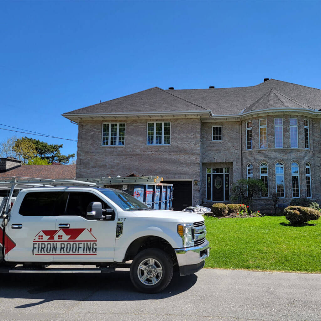 Un camion blanc Firon Roofing, desservant Ottawa / Gatineau, est stationné devant une grande maison en brique à deux étages avec un toit foncé et de hautes fenêtres, par une journée ensoleillée. Une benne et du matériel sont visibles dans l’allée.