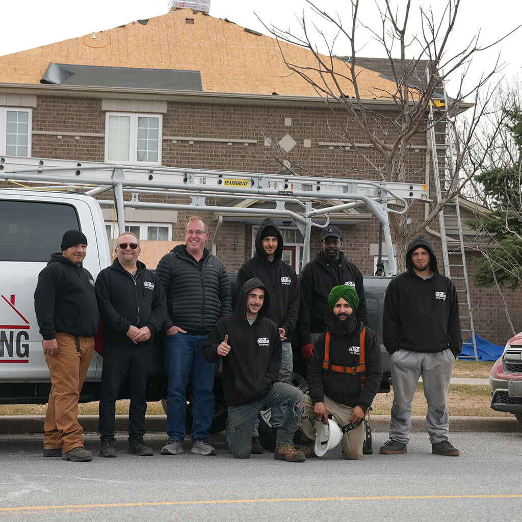 Un groupe de huit personnes, incluant des couvreurs qualifiés d’Ottawa, pose devant un camion Firon Roofing dans une rue de banlieue, avec une maison en construction derrière eux. Certains portent des vêtements de travail et des chandails à capuchon; l’ambiance est décontractée et amicale.